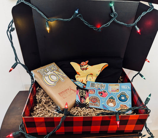 A red and black plaid gift box is surrounded with multicoloured string lights. Inside the box is a black t-shirt featuring the character rad ghost, a hard enamel pin of the character Ali and a VHS tape. The pin is in black nickel. A VHS tape is covered in craft paper. There is a sticker on it of The Big Con logo that is black, white, hot pink, yellow and blue. Below that it is stamped with the Mighty Yell logo in hot pink. There is a brightly coloured sticker sheet. 