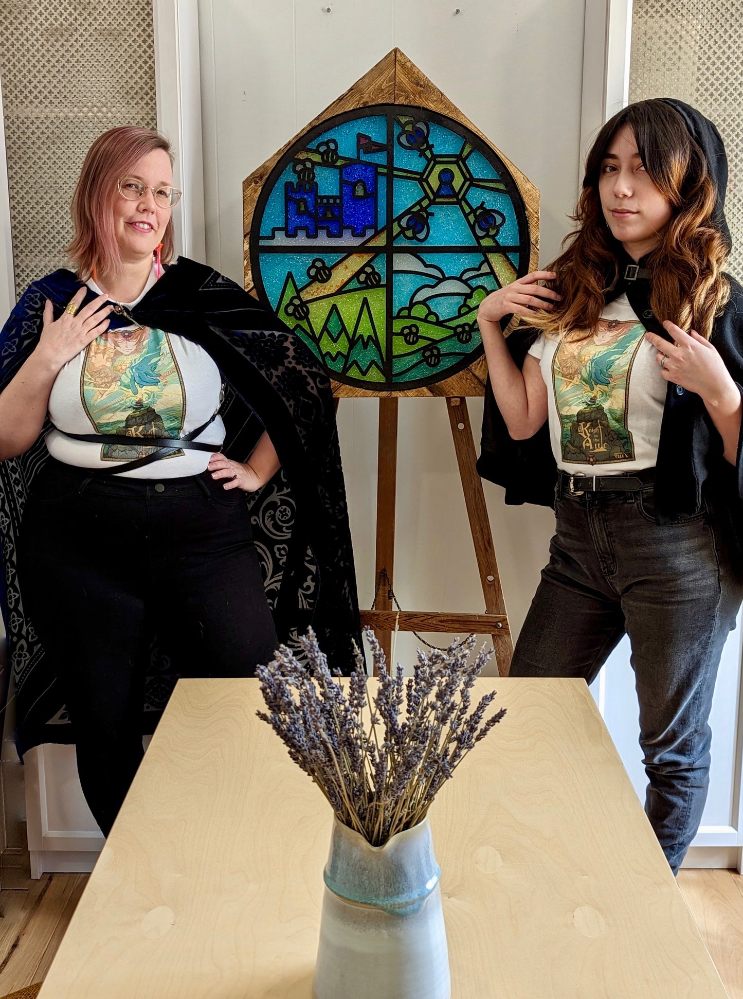 A white T-Shirt with Key Art from the game A Knight In The Attic. Featuring Characters faces and a mountain landscape in soft and vibrant blue, yellow, green, brown and peach. A character stands atop a mountain, in front of a large blue hand and a sword. In medieval style font it reads “A Knight In The Attic” and the “I” in attic is a sword. The “Mighty Yell Logo in the bottom right corner. The border is yellow with purple and burgundy flowers and green vines.