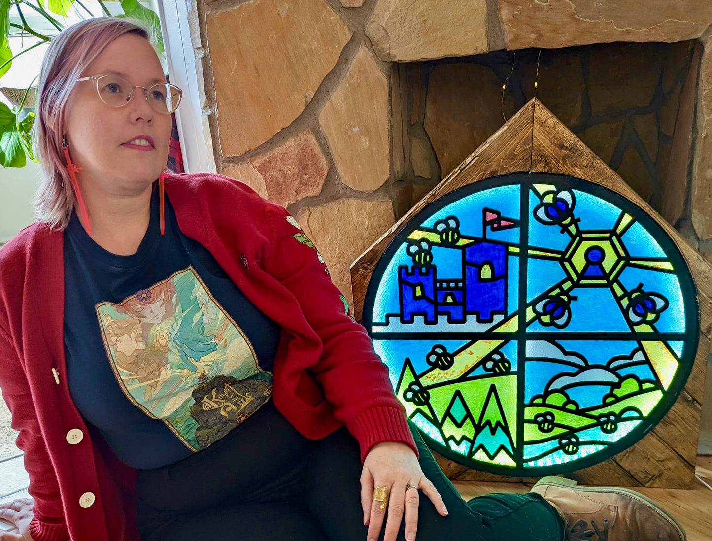 A Navy T-Shirt with Key Art from the game A Knight In The Attic. Featuring Characters faces and a mountain landscape in soft and vibrant blue, yellow, green, brown and peach. A character stands atop a mountain, in front of a large blue hand and a sword. In medieval style font it reads “A Knight In The Attic” and the “I” in attic is a sword. The “Mighty Yell Logo in the bottom right corner. The border is yellow with purple and burgundy flowers and green vines.