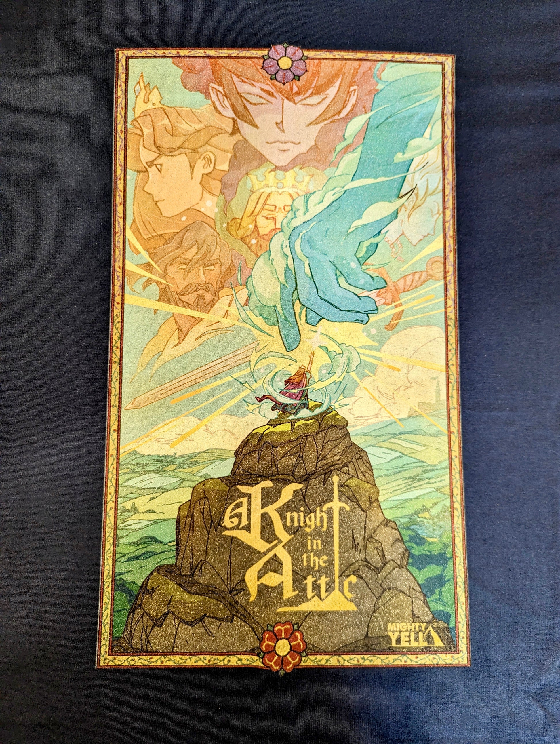 A Navy T-Shirt with Key Art from the game A Knight In The Attic. Featuring Characters faces and a mountain landscape in soft and vibrant blue, yellow, green, brown and peach. A character stands atop a mountain, in front of a large blue hand and a sword. In medieval style font it reads “A Knight In The Attic” and the “I” in attic is a sword. The “Mighty Yell Logo in the bottom right corner. The border is yellow with purple and burgundy flowers and green vines.  