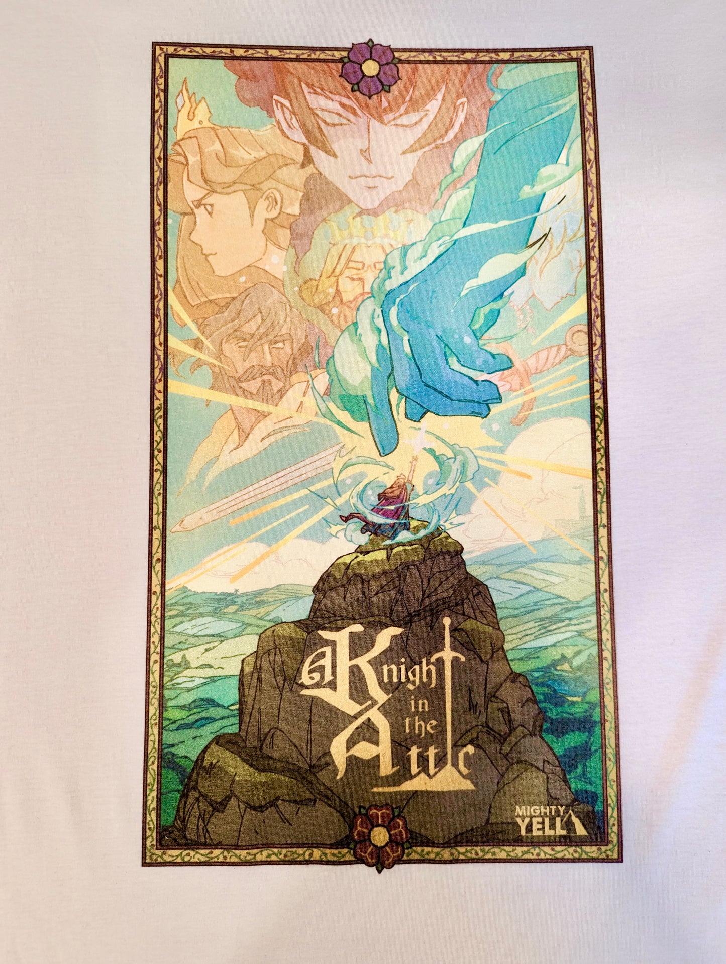 A white T-Shirt with Key Art from the game A Knight In The Attic. Featuring Characters faces and a mountain landscape in soft and vibrant blue, yellow, green, brown and peach. A character stands atop a mountain, in front of a large blue hand and a sword. In medieval style font it reads “A Knight In The Attic” and the “I” in attic is a sword. The “Mighty Yell Logo in the bottom right corner. The border is yellow with purple and burgundy flowers and green vines.