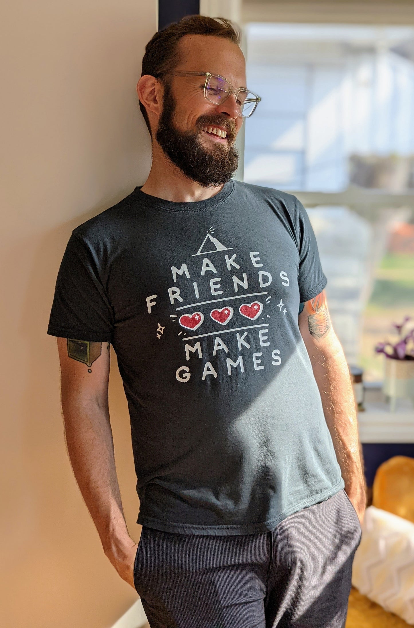 A t-shirt with the words "Make Friends Make Games," featuring 3 hearts like in a Zelda Game, and the MIghty Yell Mountain Logo, arranged aesthetically on the front of the shirt. Charcoal grey shirt with white text and red hearts.