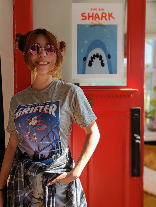 A t-shirt that says "GRIFTER," stylized like an old THRASHER magazine cover, featuring Ali from The Big Con jumping on a skateboard over some stairs. Light heathered grey shirt, with the image stylized like a magazine cover in rectangle on the front of the shirt. Magazine cover colours are largely Blue, light blue, and pink, like a stylized night sky. The magazine cover has a distressed look, like an old print.