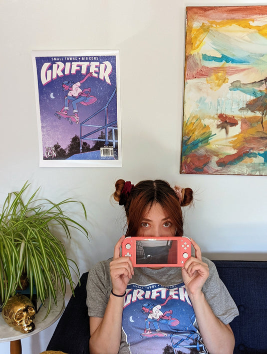 A poster that says "GRIFTER," stylized like an old THRASHER magazine cover, featuring Ali from The Big Con jumping on a skateboard over some stairs. Image stylized like a magazine cover in rectangle on the front of the shirt. Magazine cover colours are largely Blue, light blue, and pink, like a stylized night sky. The magazine cover has a distressed look, like an old print.