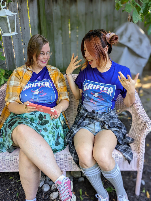 A t-shirt that says "GRIFTER," stylized like an old THRASHER magazine cover, featuring Ali from The Big Con jumping on a skateboard over some stairs. Deep purple shirt, with the image stylized like a magazine cover in rectangle on the front of the shirt. Magazine cover colours are largely Blue, light blue, and pink, like a stylized night sky. The magazine cover has a distressed look, like an old print.
