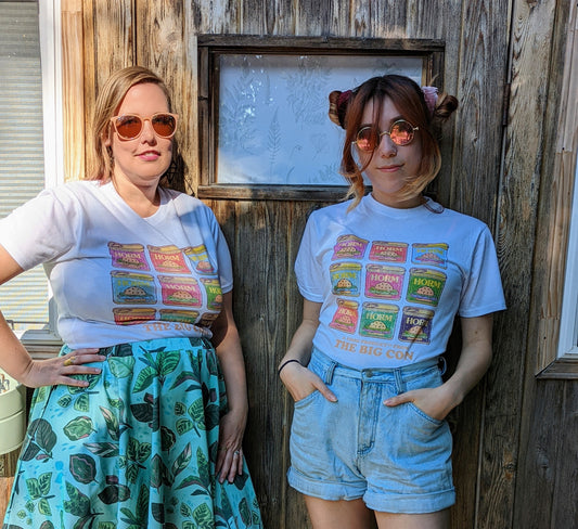 A white t-hirt with nine HORM cans, that parody the canned meat SPAM. The cans are inspired by the soup can print by Andy Warhol. They are placed in a 3 x 3 grid and are all different colours including purple, red, light blue, medium blue, bright pink, green, rose, bright green and dark purple. Below the cans it says “A FOOD PRODUCT FROM THE BIG CON”. After the word “PRODUCT” there is a trademark symbol.