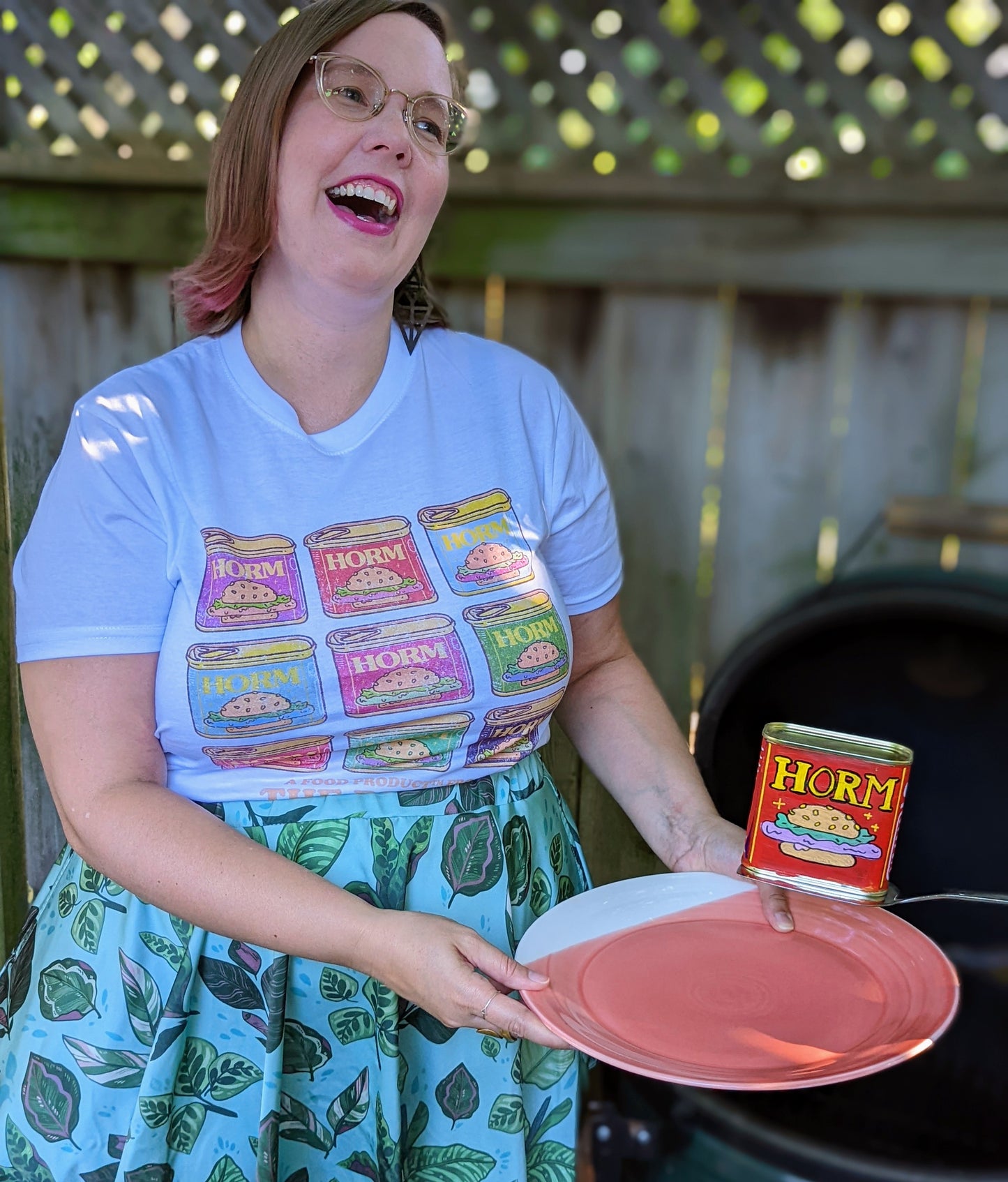 A white t-hirt with nine HORM cans, that parody the canned meat SPAM. The cans are inspired by the soup can print by Andy Warhol. They are placed in a 3 x 3 grid and are all different colours including purple, red, light blue, medium blue, bright pink, green, rose, bright green and dark purple. Below the cans it says “A FOOD PRODUCT FROM THE BIG CON”. After the word “PRODUCT” there is a trademark symbol.
