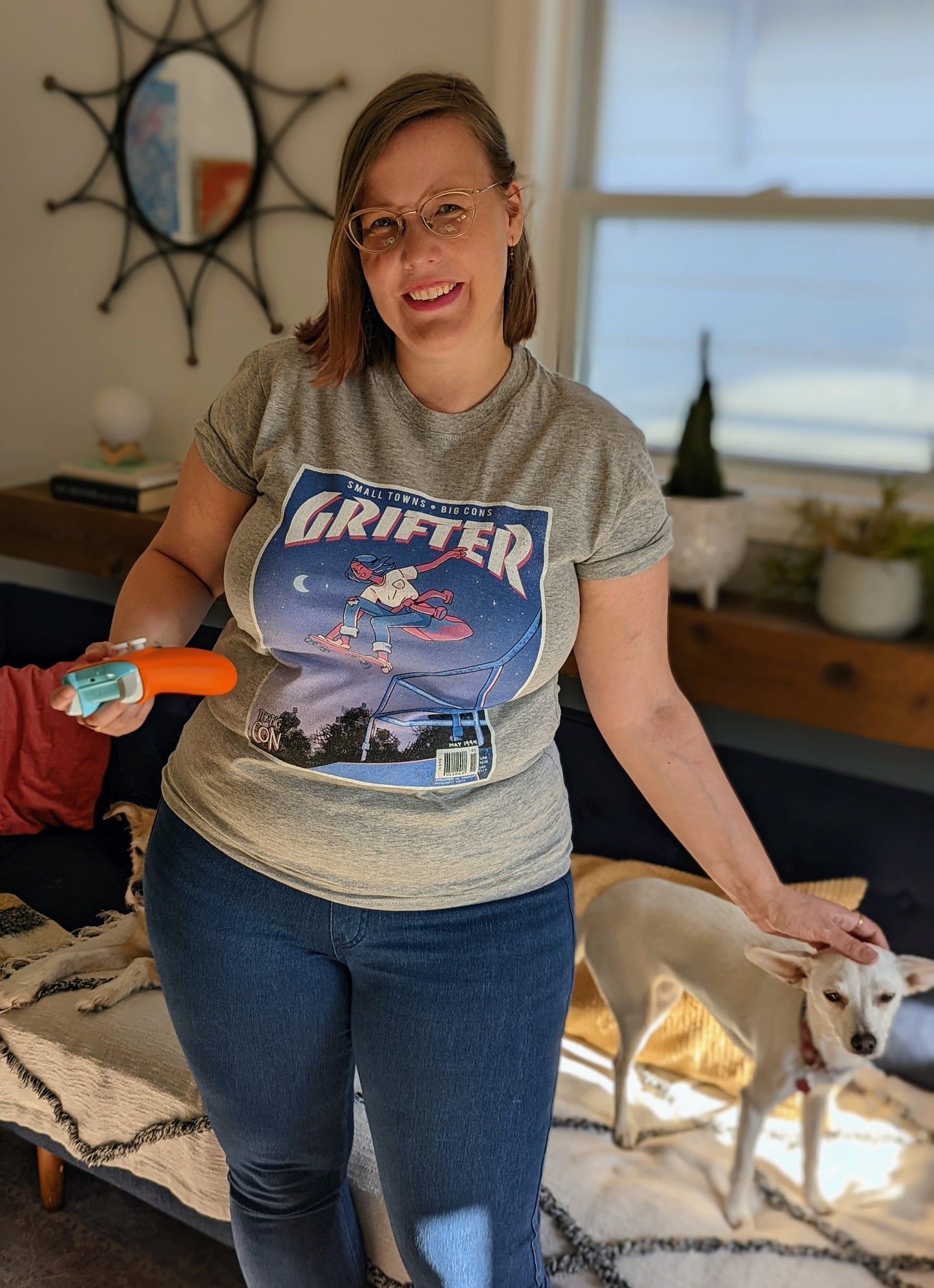 A t-shirt that says "GRIFTER," stylized like an old THRASHER magazine cover, featuring Ali from The Big Con jumping on a skateboard over some stairs. Light heathered grey shirt, with the image stylized like a magazine cover in rectangle on the front of the shirt. Magazine cover colours are largely Blue, light blue, and pink, like a stylized night sky. The magazine cover has a distressed look, like an old print.