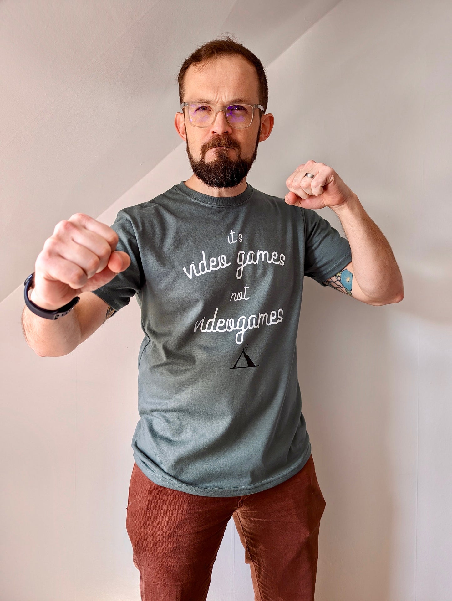 A Sage Green T-shirt that has cursive writing on it in white. In lowercase letters the first line reads “it’s”, the second line “video games” using two words, the third line “not” and the fourth line “videogames” spelt as one word. In the centre below the text is the Mighty Yell logo in black which features the illustration of a mountain and a person on top yelling. There are three small lines coming out of the person’s mouth.