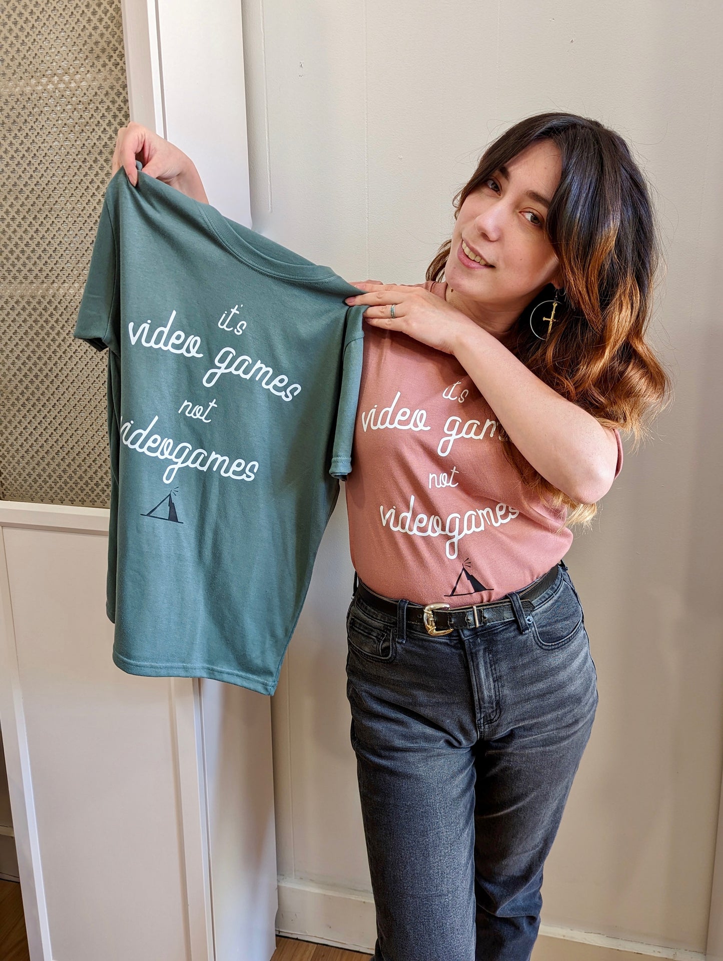A Sage Green T-shirt and a Dusty Rose T-Shirt, both have cursive writing on it in white. In lowercase letters the first line reads “it’s”, the second line “video games” using two words, the third line “not” and the fourth line “videogames” spelt as one word. In the centre below the text is the Mighty Yell logo in black which features the illustration of a mountain and a person on top yelling. There are three small lines coming out of the person’s mouth.