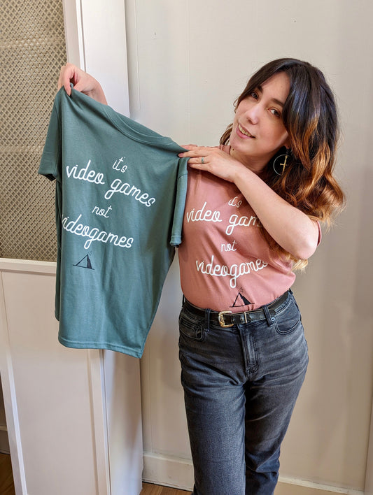 A Sage Green T-shirt and a Dusty Rose T-Shirt, both have cursive writing on it in white. In lowercase letters the first line reads “it’s”, the second line “video games” using two words, the third line “not” and the fourth line “videogames” spelt as one word. In the centre below the text is the Mighty Yell logo in black which features the illustration of a mountain and a person on top yelling. There are three small lines coming out of the person’s mouth. 