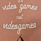 A Dusty Rose T-shirt that has cursive writing on it in white. In lowercase letters the first line reads “it’s”, the second line “video games” using two words, the third line “not” and the fourth line “videogames” spelt as one word. In the centre below the text is the Mighty Yell logo in black which features the illustration of a mountain and a person on top yelling. There are three small lines coming out of the person’s mouth.