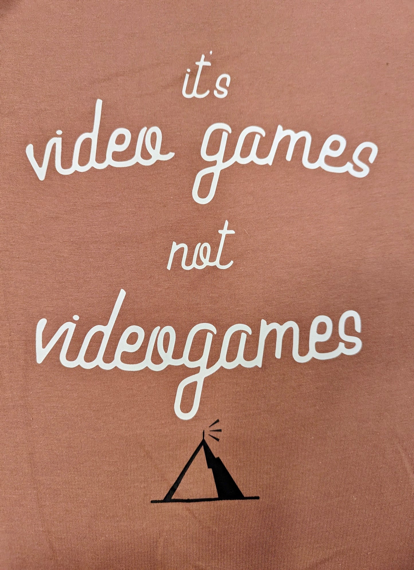A Dusty Rose T-shirt that has cursive writing on it in white. In lowercase letters the first line reads “it’s”, the second line “video games” using two words, the third line “not” and the fourth line “videogames” spelt as one word. In the centre below the text is the Mighty Yell logo in black which features the illustration of a mountain and a person on top yelling. There are three small lines coming out of the person’s mouth.