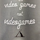 A Sage Green T-Shirt, with cursive writing on it in white. In lowercase letters the first line reads “it’s”, the second line “video games” using two words, the third line “not” and the fourth line “videogames” spelt as one word. In the centre below the text is the Mighty Yell logo in black which features the illustration of a mountain and a person on top yelling. There are three small lines coming out of the person’s mouth. 