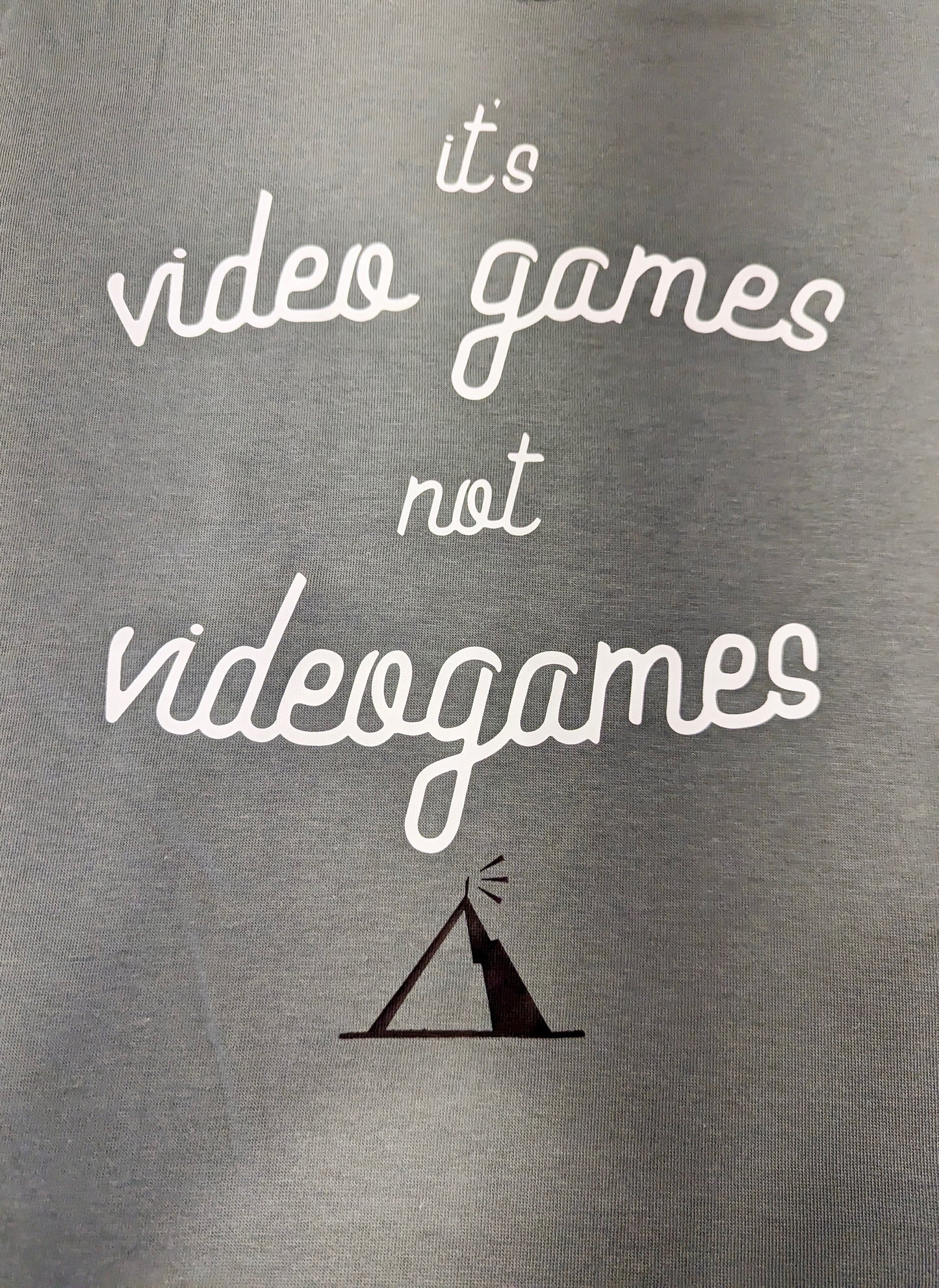 A Sage Green T-Shirt, with cursive writing on it in white. In lowercase letters the first line reads “it’s”, the second line “video games” using two words, the third line “not” and the fourth line “videogames” spelt as one word. In the centre below the text is the Mighty Yell logo in black which features the illustration of a mountain and a person on top yelling. There are three small lines coming out of the person’s mouth.