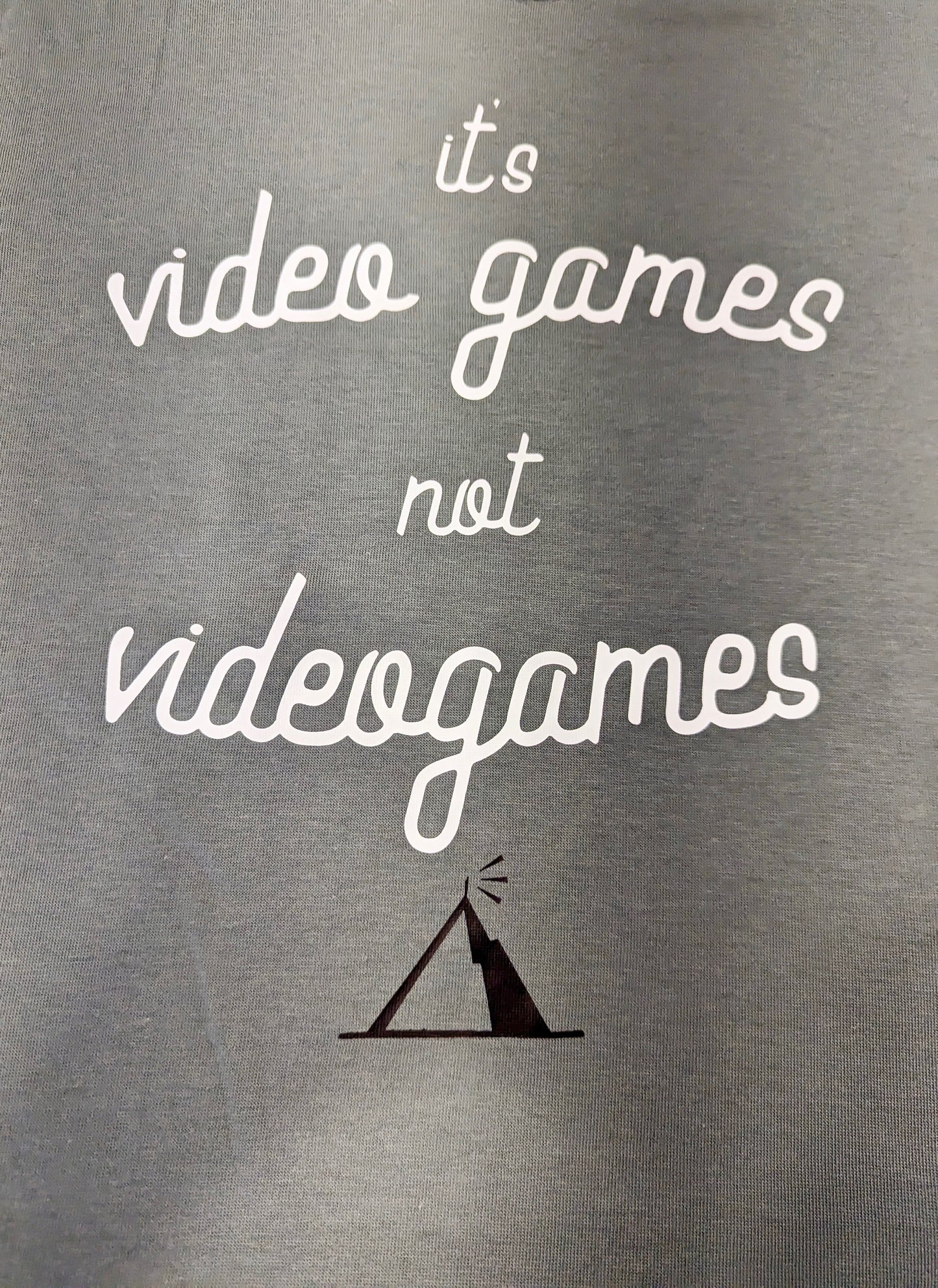 A Sage Green T-Shirt, with cursive writing on it in white. In lowercase letters the first line reads “it’s”, the second line “video games” using two words, the third line “not” and the fourth line “videogames” spelt as one word. In the centre below the text is the Mighty Yell logo in black which features the illustration of a mountain and a person on top yelling. There are three small lines coming out of the person’s mouth. 