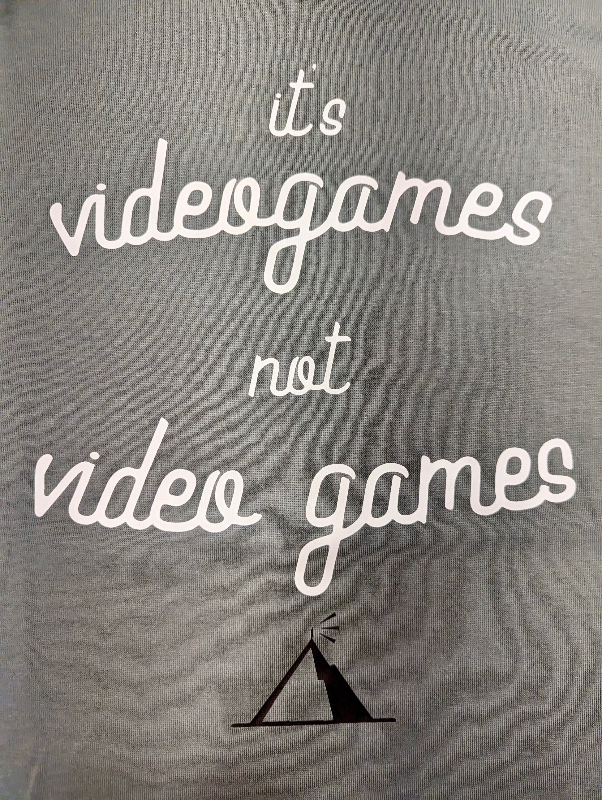 A Sage Green T-Shirt, with cursive writing on it in white. In lowercase letters the first line reads “it’s”, the second line “videogames” spelt as one word, the third line “not” and the fourth line “video games” using two words. In the centre below the text is the Mighty Yell logo in black which features the illustration of a mountain and a person on top yelling. There are three small lines coming out of the person’s mouth.