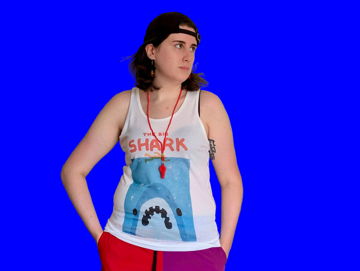 A person wearing a white tank top white tank top stylized to parody the movie Jaws. It has with a blue shark graphic and the text 'THE BIG SHARK' in red, paired with red and purple pants, standing in front of a bright blue background.