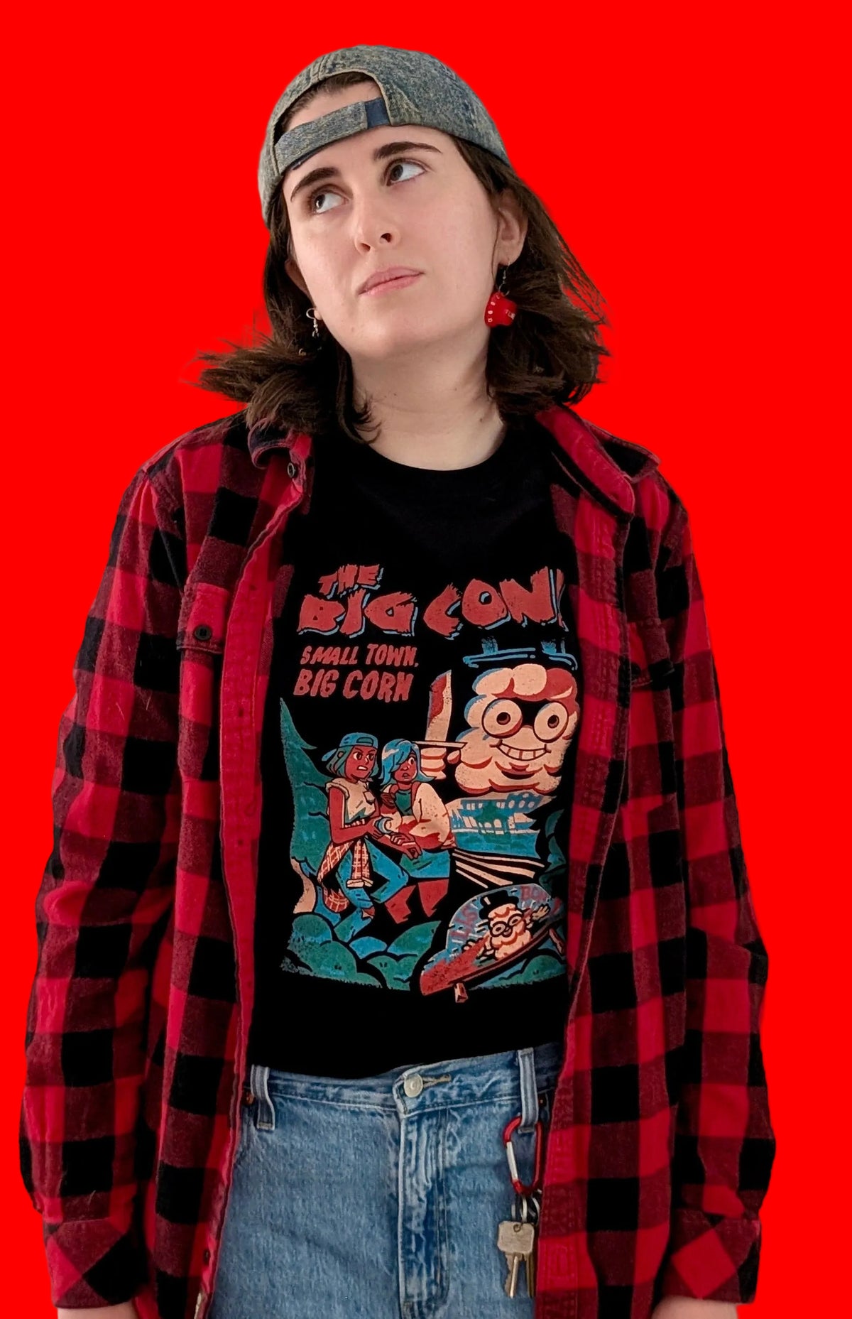 A person standing against a solid red background, wearing black graphic T-shirts featuring a characters Ali and Mari from the game The Big Con, hiding from an enormous cob of corn holding a knife.  The text in red says 'THE BIG CON! A SMALL TOWN BIG CORN.' 