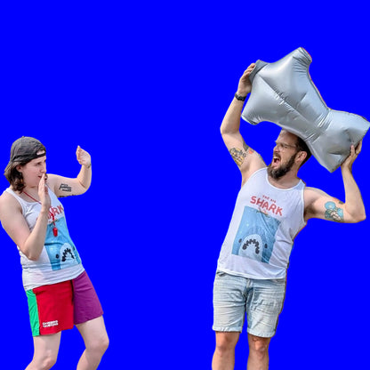 Two people wearing matching white tank tops stylized to parody the movie Jaws. At the top of the shirt it says “THE BIG SHARK” in a cartoonish font in red. Below that is a cartoon outline of a person swimming over of an enormous shark mouth. The shark is blue, white and black and it sits on a distressed, grainy, light blue square background. They are posing against a solid bright blue background. The person on the right holds a large silver inflatable mannequin torso above their head.