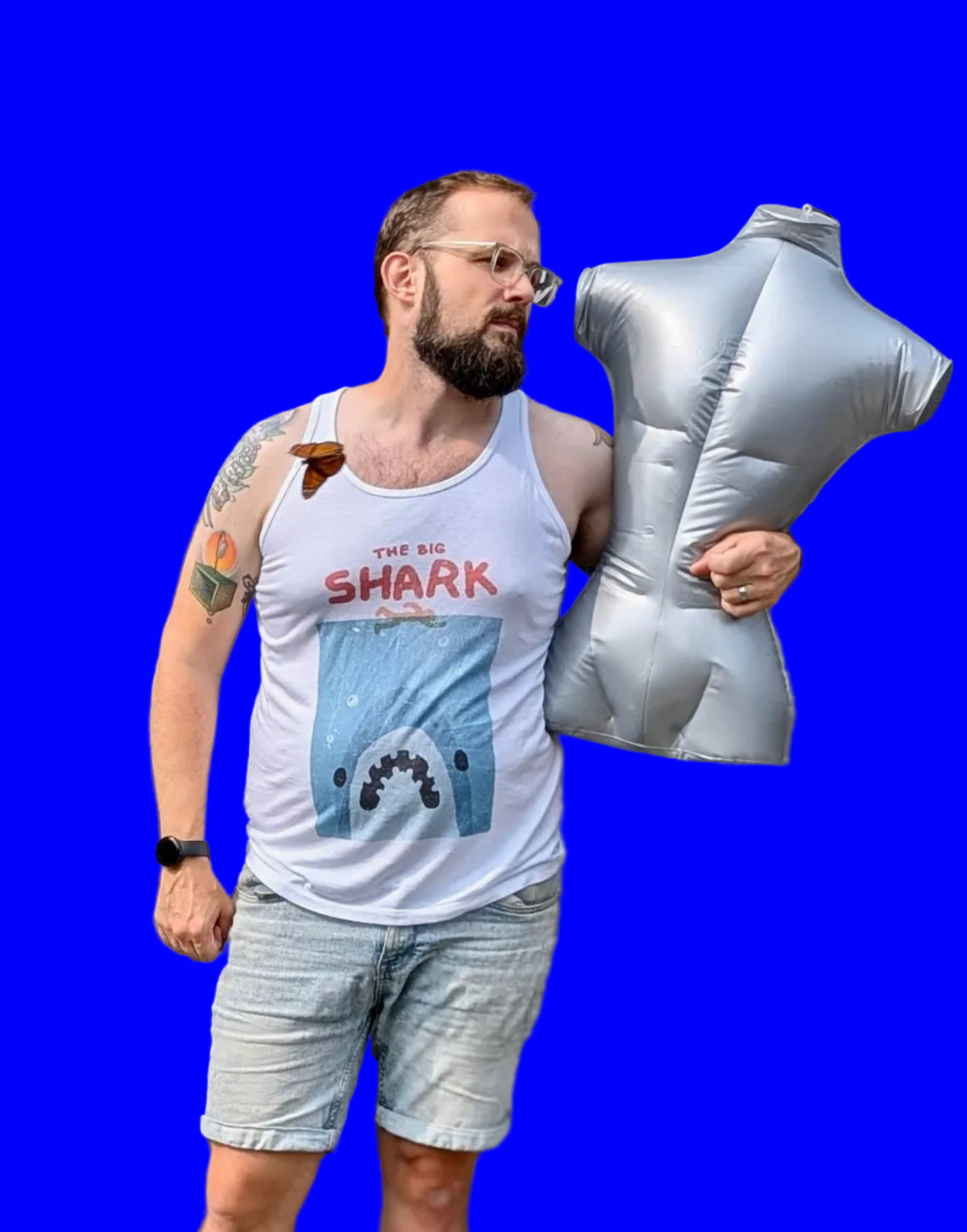 A man wearing a white tank tops stylized to parody the movie Jaws. At the top of the shirt it says “THE BIG SHARK” in a cartoonish font in red. Below that is a cartoon outline of a person swimming over of an enormous shark mouth. The shark is blue, white and black and it sits on a distressed, grainy, light blue square background. The person on the right holds a large silver inflatable mannequin torso to his side against a bright blue background.