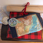 A red and black plaid gift box. On top of it sits a navy t-shirt featuring the key art from A Knight In The Attic. Featuring characters faces and a mountain landscape in soft and vibrant blue, yellow, green, brown and peach. A character stands atop a mountain, in front of a large blue hand and a sword. To the left of the the t-shirt is a round transparent sticker in the style of stained glass in yellow, green, blue and red. At the top is a brown poster tube tied with a thin red velvet ribbon.