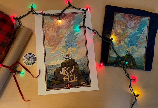 On a table sits a navy t-shirt featuring the key art from A Knight In The Attic. Characters faces and a mountain landscape in blue, yellow, green, brown and peach. A character stands atop a mountain, in front of a large blue hand and a sword. To the left of  the t-shirt is a poster with the same key art. In between is a round transparent sticker in the style of stained glass in yellow, green, blue and red.