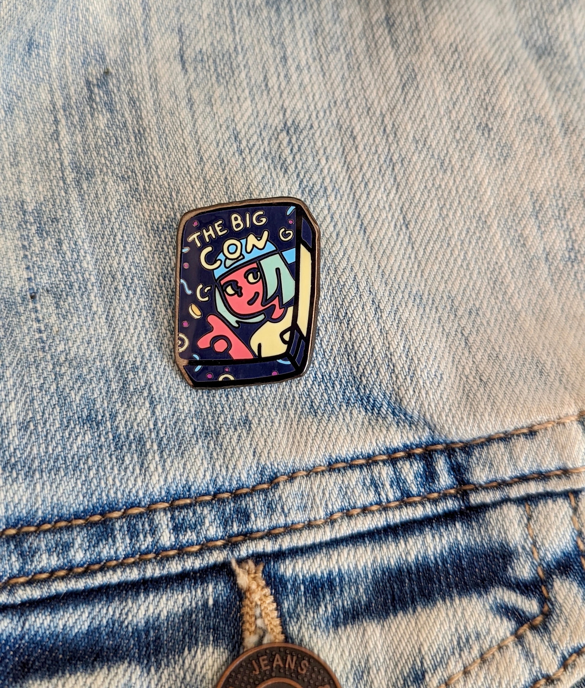 A hard enamel pin in black nickel. The pin measures 1.5”. It is of Ali, the main character from The Big Con, as if she is on the cover of a VHS tape. She is wearing a blue backwards baseball cap and a yellow shirt. Through the pin there are curved shapes in pink, blue and yellow. Ali is flipping a gold coin in the air.  