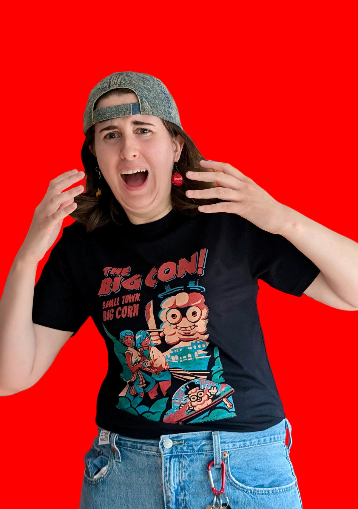 A person standing against a solid red background, wearing black graphic T-shirts featuring a characters Ali and Mari from the game The Big Con, hiding from an enormous cob of corn holding a knife.  The text in red says 'THE BIG CON! A SMALL TOWN BIG CORN.' 