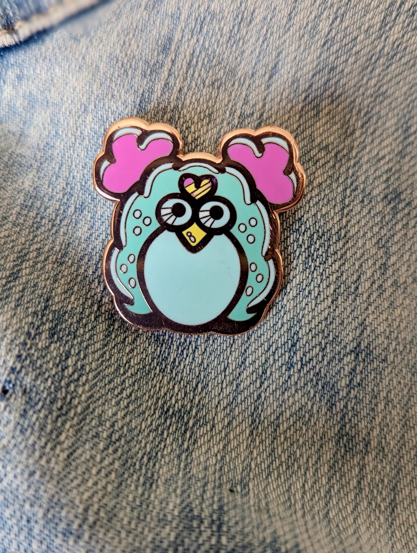 A hard enamel pin in rose gold. The pin measures 1.5”.  It is of a 90’s inspired collectable plush toy in light pink and turquoise.