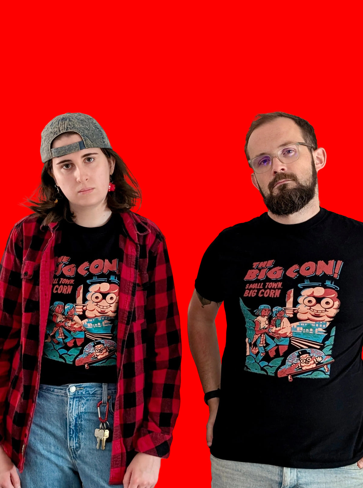 Two people standing against a solid red background, both wearing black graphic T-shirts featuring a characters Ali and Mari from the game The Big Con, hiding from an enormous cob of corn holding a knife.  The text in red says 'THE BIG CON! A SMALL TOWN BIG CORN.' The person on the left also wears a red-and-black flannel shirt, jeans, and a grey hat, while the person on the right wears light grey jeans.