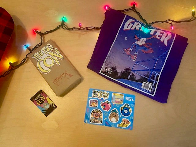 Unboxed items sit on a table. A purple grifter style t-shirt, a hard enamel pin of a pink and turqoise toy in the style of a 90s collectible toy. The pin is in rose gold. A VHS tape is covered in craft paper. There is a sticker on it of The Big Con logo that is black, white, hot pink, yellow and blue. Below that it is stamped with the Mighty Yell logo in hot pink. There is a brightly coloured sticker sheet featuring eight stickers from The Big Con.