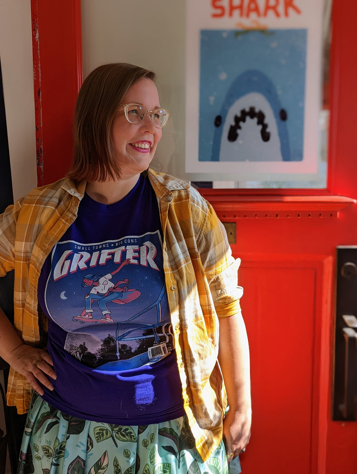 A t-shirt that says "GRIFTER," stylized like an old THRASHER magazine cover, featuring Ali from The Big Con jumping on a skateboard over some stairs. Deep purple shirt, with the image stylized like a magazine cover in rectangle on the front of the shirt. Magazine cover colours are largely Blue, light blue, and pink, like a stylized night sky. The magazine cover has a distressed look, like an old print.