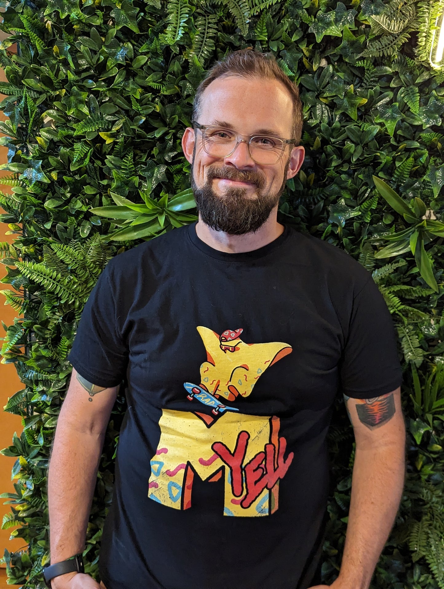 A person standing in front of a plant wall wearing a black shirt with the character Rad Ghost skateboarding over top a large "M" and the word "yell" in the style of MTV. The images are in yellow with pops of Orange, blue and pink.