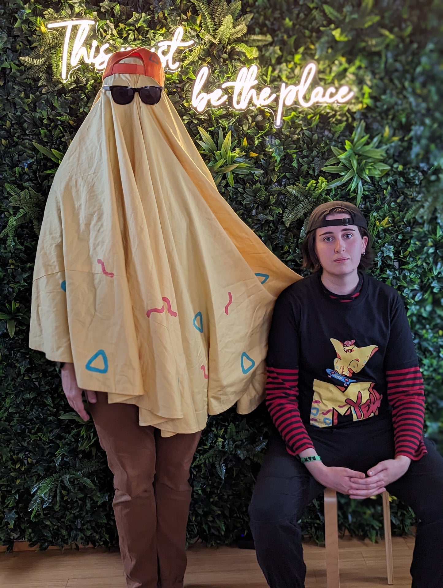 A ghost wearing a yellow sheet with an orange baseball cap, and cool mirrored sunglasses stands in front of a plant wall beside a person sitting on a stool wearing a black shirt with the character Rad Ghost skateboarding over top a large "M" and the word "yell" in the style of MTV. The images are in yellow with pops of Orange, blue and pink.