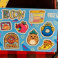 A brightly coloured sticker sheet for the big con, with a blue background, and several stickers outlined on it that can be peeled off: a worm in an apple, a can of horm, a man holding a magic 8ball that says bro, a coconut burblo a slice of pizza wearing sunglasses, a logo for mallton mall, and a sticker of rad ghost with the word rad.