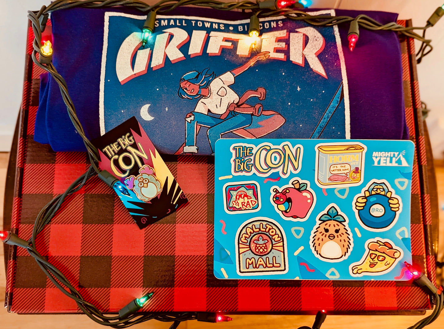A red and black plaid gift box is surrounded with multicoloured string lights. Inside the box is a purple grifter style t-shirt, a hard enamel pin of a pink and turqoise toy in the style of a 90s collectible toy. The pin is in rose gold. There is a brightly coloured sticker sheet featuring eight stickers from The Big Con.