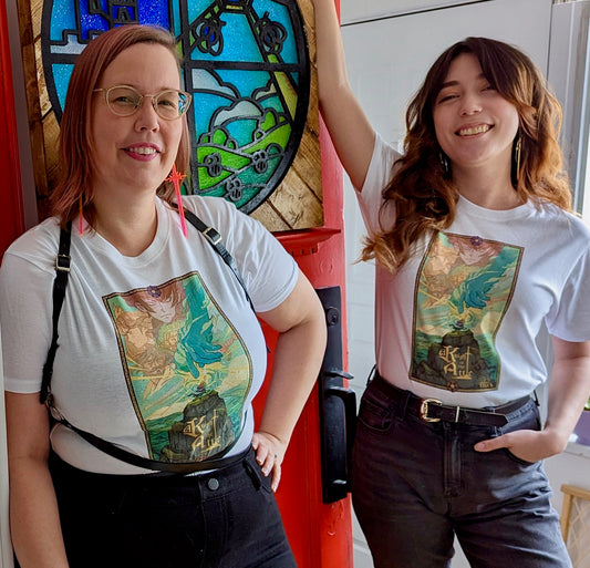 A white T-Shirt with Key Art from the game A Knight In The Attic. Featuring Characters faces and a mountain landscape in soft and vibrant blue, yellow, green, brown and peach. A character stands atop a mountain, in front of a large blue hand and a sword. In medieval style font it reads “A Knight In The Attic” and the “I” in attic is a sword. The “Mighty Yell Logo in the bottom right corner. The border is yellow with purple and burgundy flowers and green vines.  