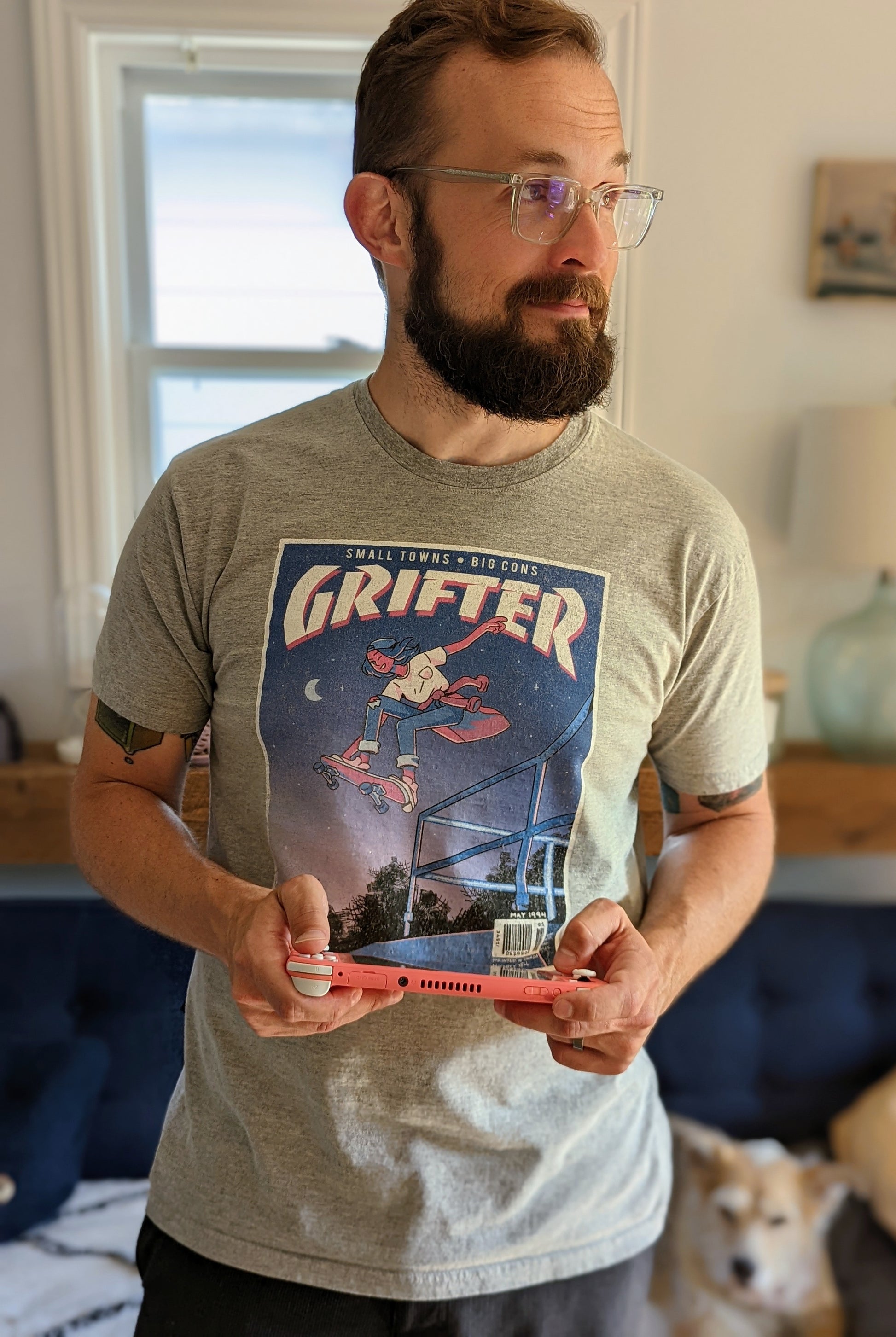 A t-shirt that says "GRIFTER," stylized like an old THRASHER magazine cover, featuring Ali from The Big Con jumping on a skateboard over some stairs. Light heathered grey shirt, with the image stylized like a magazine cover in rectangle on the front of the shirt. Magazine cover colours are largely Blue, light blue, and pink, like a stylized night sky. The magazine cover has a distressed look, like an old print.