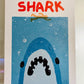 A poster stylized to parody the movie Jaws. At the top it says “THE BIG SHARK” in a cartoonish font in red. Below that is a cartoon outline of a person swimming over of an enormous Shark mouth. The shark is blue, white and black and it sits on a distressed, grainy, light blue square background. In the bottom right corner there is small blue THE BIG CON logo. The whole poster sits on a white background.