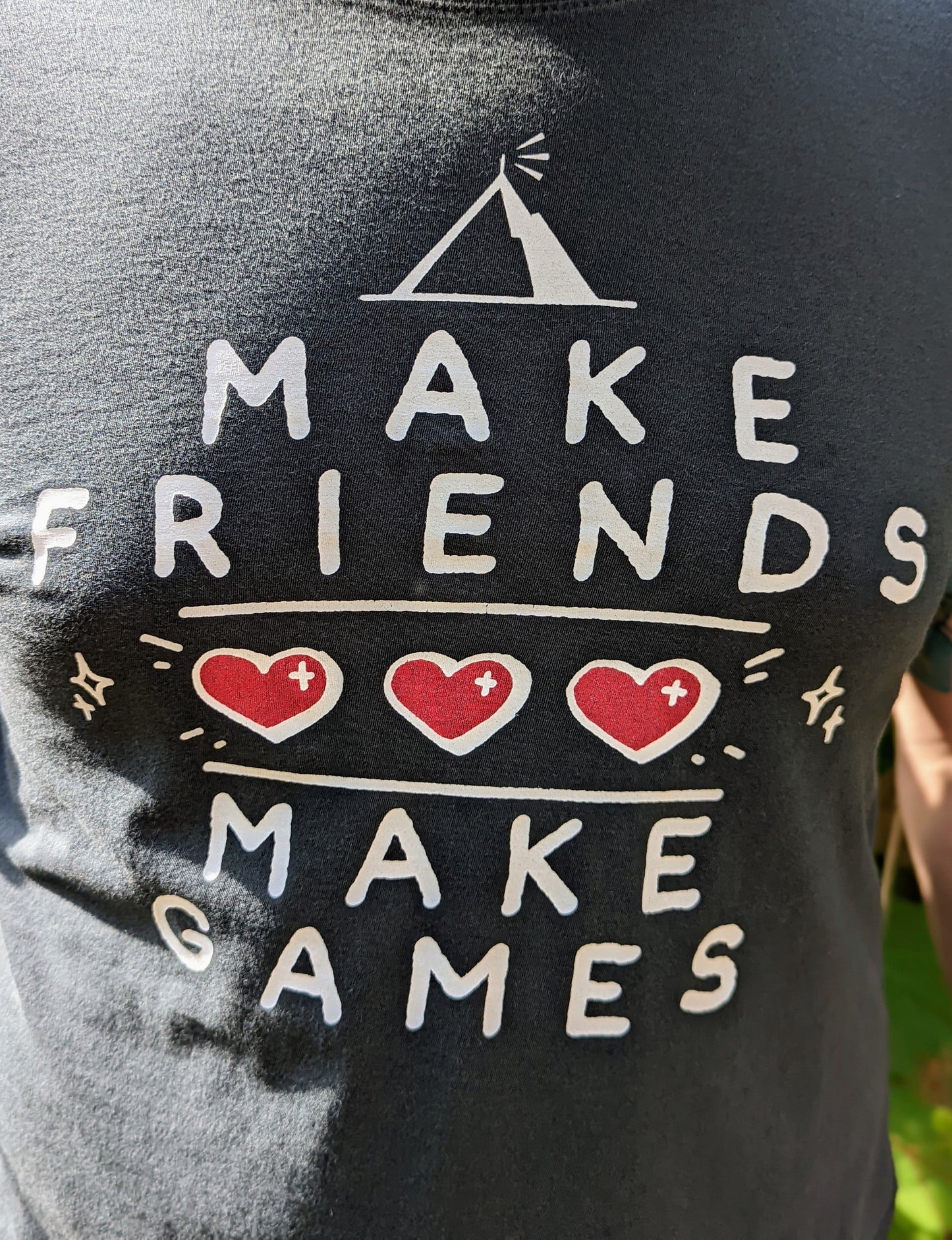 A t-shirt with the words "Make Friends Make Games," featuring 3 hearts like in a Zelda Game, and the MIghty Yell Mountain Logo, arranged aesthetically on the front of the shirt. Charcoal grey shirt with white text and red hearts.