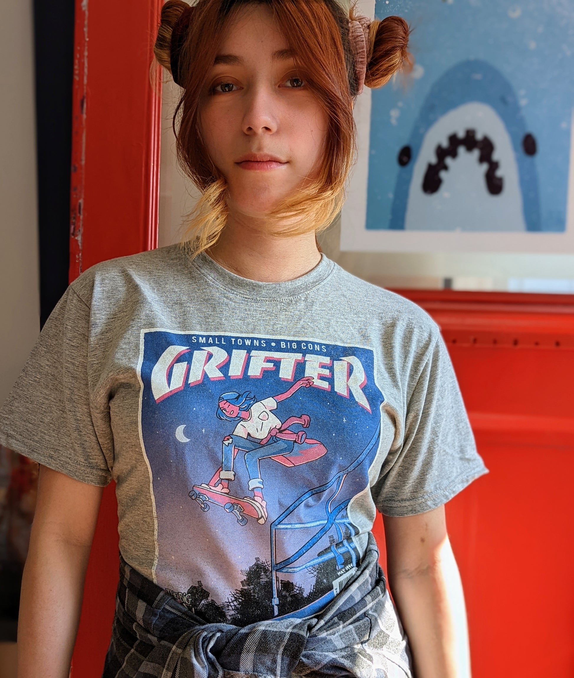 A t-shirt that says "GRIFTER," stylized like an old THRASHER magazine cover, featuring Ali from The Big Con jumping on a skateboard over some stairs. Light heathered grey shirt, with the image stylized like a magazine cover in rectangle on the front of the shirt. Magazine cover colours are largely Blue, light blue, and pink, like a stylized night sky. The magazine cover has a distressed look, like an old print.