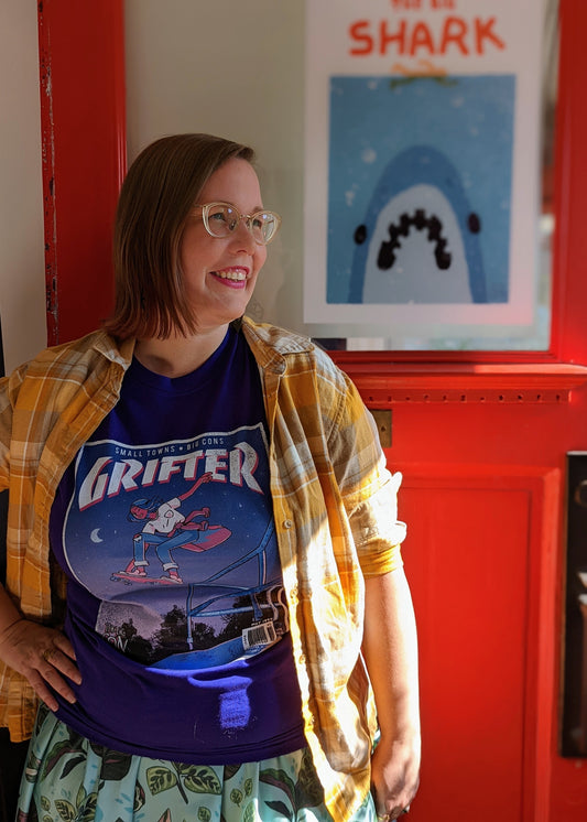A t-shirt that says "GRIFTER," stylized like an old THRASHER magazine cover, featuring Ali from The Big Con jumping on a skateboard over some stairs. Deep purple shirt, with the image stylized like a magazine cover in rectangle on the front of the shirt. Magazine cover colours are largely Blue, light blue, and pink, like a stylized night sky. The magazine cover has a distressed look, like an old print.