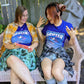 A t-shirt that says "GRIFTER," stylized like an old THRASHER magazine cover, featuring Ali from The Big Con jumping on a skateboard over some stairs. Deep purple shirt, with the image stylized like a magazine cover in rectangle on the front of the shirt. Magazine cover colours are largely Blue, light blue, and pink, like a stylized night sky. The magazine cover has a distressed look, like an old print.