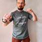 A Sage Green T-shirt that has cursive writing on it in white. In lowercase letters the first line reads “it’s”, the second line “video games” using two words, the third line “not” and the fourth line “videogames” spelt as one word. In the centre below the text is the Mighty Yell logo in black which features the illustration of a mountain and a person on top yelling. There are three small lines coming out of the person’s mouth. 