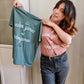 A Sage Green T-shirt and a Dusty Rose T-Shirt, both have cursive writing on it in white. In lowercase letters the first line reads “it’s”, the second line “video games” using two words, the third line “not” and the fourth line “videogames” spelt as one word. In the centre below the text is the Mighty Yell logo in black which features the illustration of a mountain and a person on top yelling. There are three small lines coming out of the person’s mouth. 