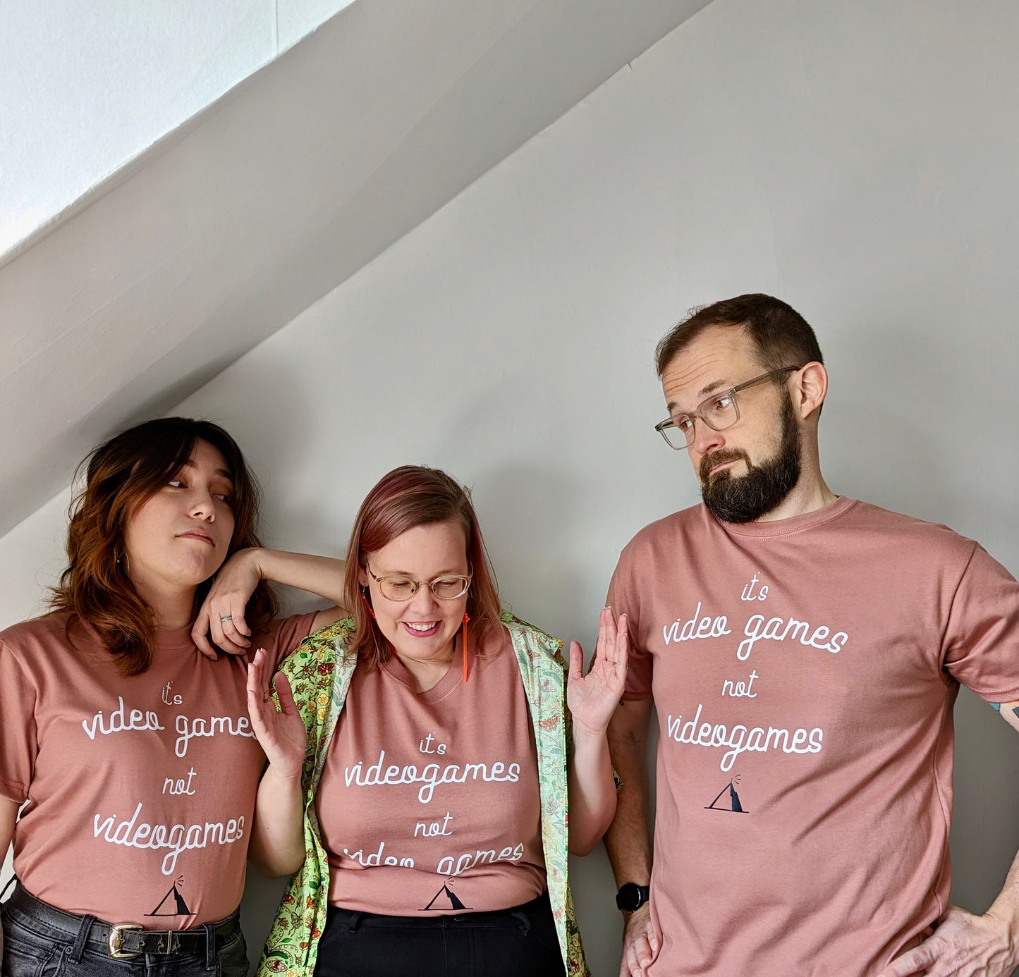 A Dusty Rose T-shirt that has cursive writing on it in white. In lowercase letters the first line reads “it’s”, the second line “video games” using two words, the third line “not” and the fourth line “videogames” spelt as one word. In the centre below the text is the Mighty Yell logo in black which features the illustration of a mountain and a person on top yelling. There are three small lines coming out of the person’s mouth.