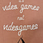 A Dusty Rose T-shirt that has cursive writing on it in white. In lowercase letters the first line reads “it’s”, the second line “video games” using two words, the third line “not” and the fourth line “videogames” spelt as one word. In the centre below the text is the Mighty Yell logo in black which features the illustration of a mountain and a person on top yelling. There are three small lines coming out of the person’s mouth.
