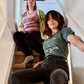 A Dusty Rose T-Shirt and a Sage Green T-Shirt, both have cursive writing on it in white. In lowercase letters the first line reads “it’s”, the second line “videogames” spelt as one word, the third line “not” and the fourth line “video games” using two words. In the centre below the text is the Mighty Yell logo in black which features the illustration of a mountain and a person on top yelling. There are three small lines coming out of the person’s mouth.