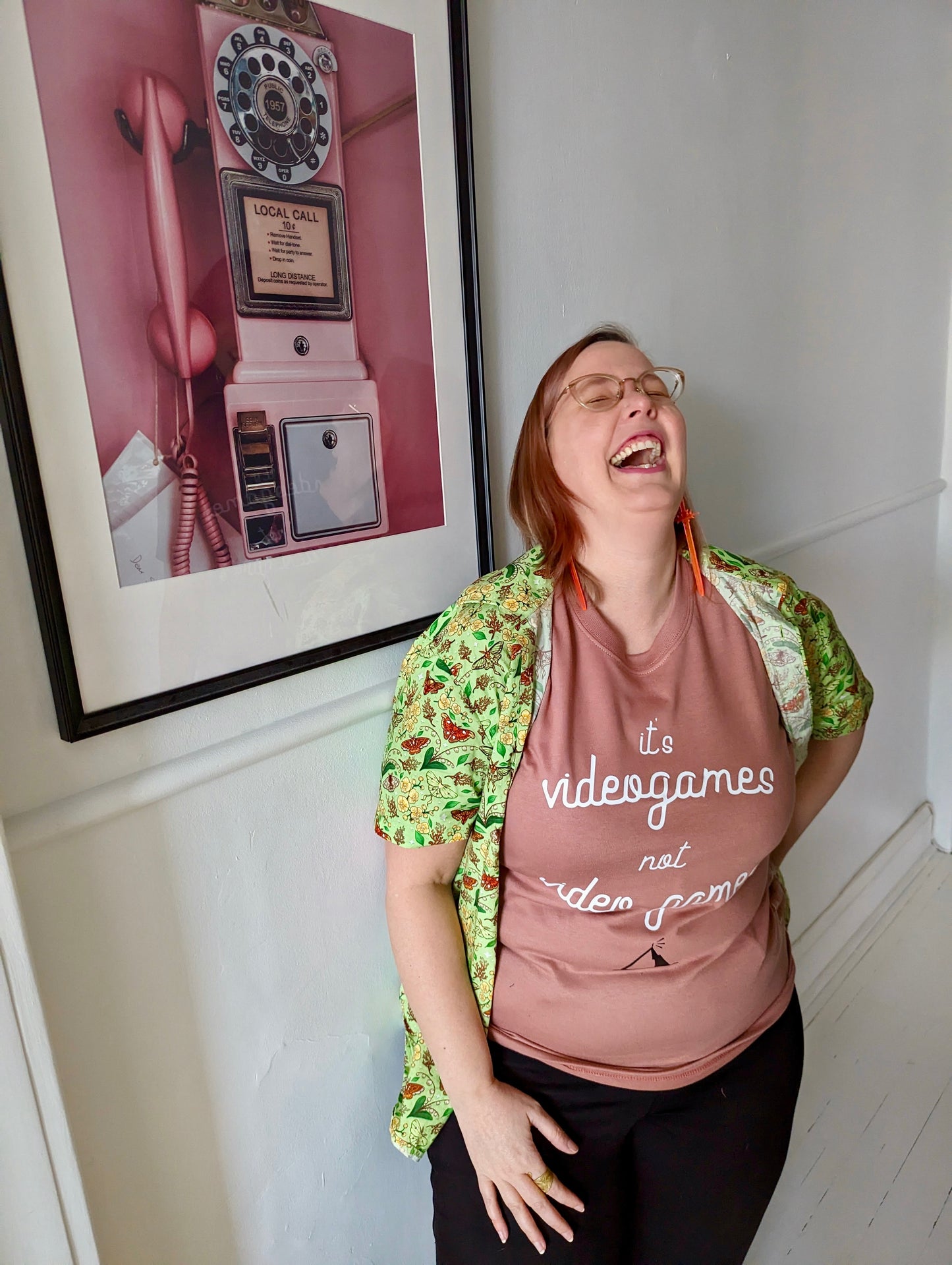 A Dusty Rose T-Shirt, with cursive writing on it in white. In lowercase letters the first line reads “it’s”, the second line “videogames” spelt as one word, the third line “not” and the fourth line “video games” using two words. In the centre below the text is the Mighty Yell logo in black which features the illustration of a mountain and a person on top yelling. There are three small lines coming out of the person’s mouth.